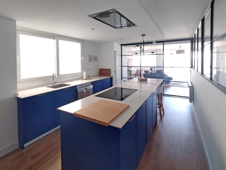 Cocina en laca mate color azul (carta NCS) con encimera compacta de resina blanca y abierta al salón separada por cristalera en forjado negro