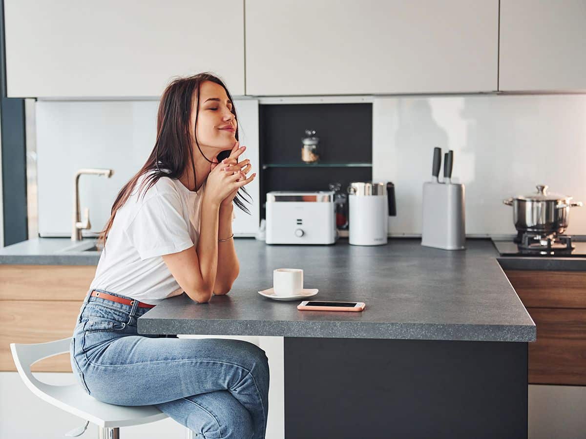 Chica joven sentada en su cocina moderna de estilo península, disfrutando de un café con los ojos cerrados, los muebles son blancos con detalles en madera y la encimera es gris.