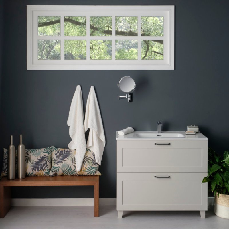 Vista frontal de mueble de lavabo en color gris claro con dos cajones con espejo pequeño extensible sobre pared, toallas colgadas en pared y pequeño banco de madera con cojines florales