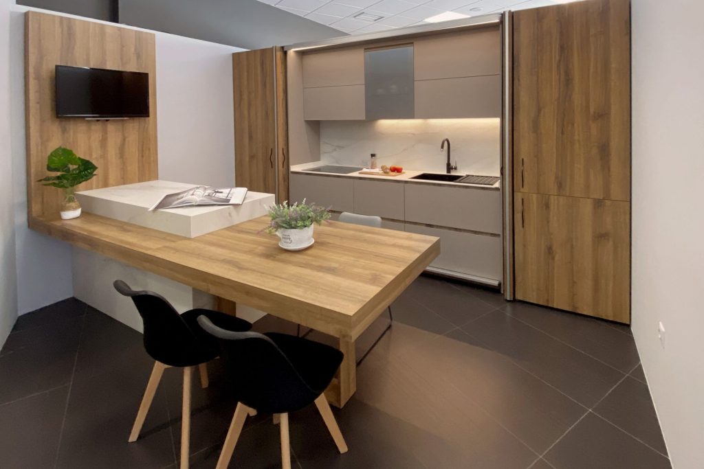 Vista lateral de cocina en blanco y madera con península con mesa integrada para comidas y TV sobre pared