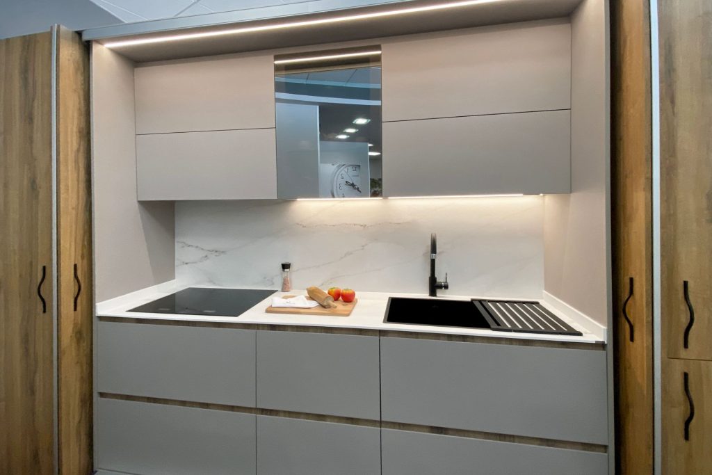 Vista frontal de cocina blanca pequeña con vitrocerámica y fregadero negro integrados, espejo entre los muebles altos y luz led bajo estos
