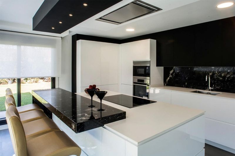 Vista lateral de cocina en combinación de blanco brillo y negro marmolado con península con vitrocerámica integrada y barra para comidas en acabado marmolado negro