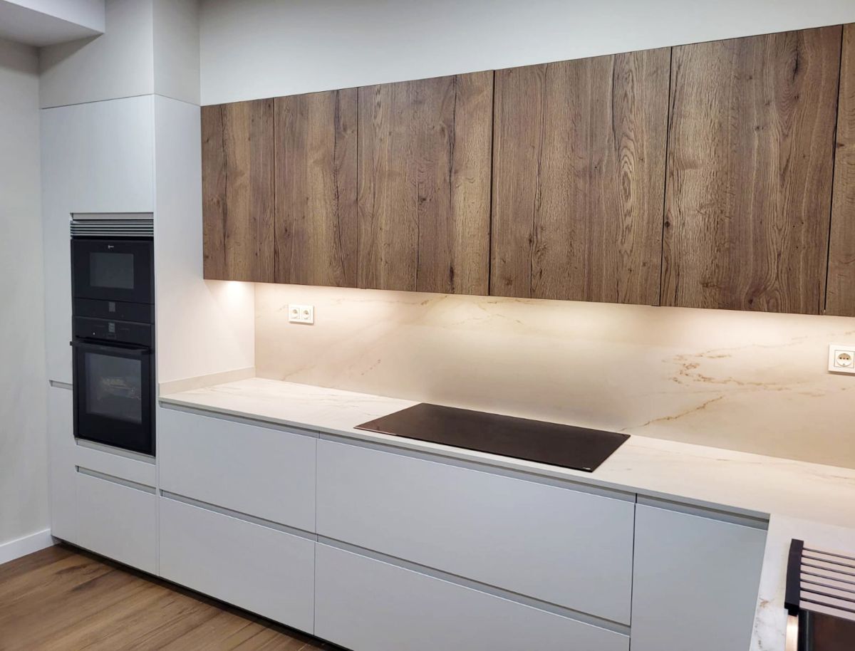 Vista lateral de cocina blanca con detalles en madera con vitrocerámica, electrodomésticos integrados y luz bajo los muebles