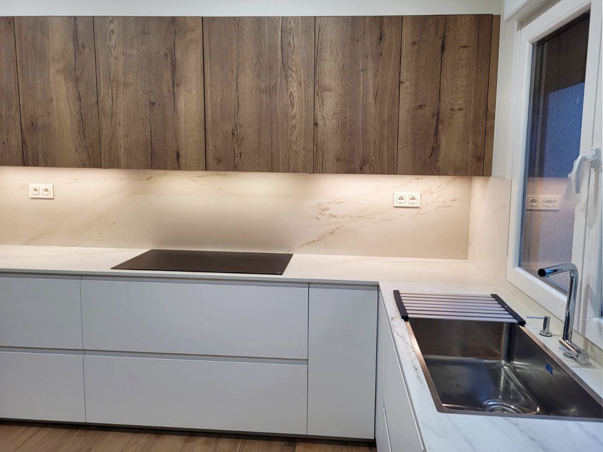 Vista frontal de cocina blanca en ele con muebles altos de madera, encimera blanca con vitrocerámica y fregadero integrado