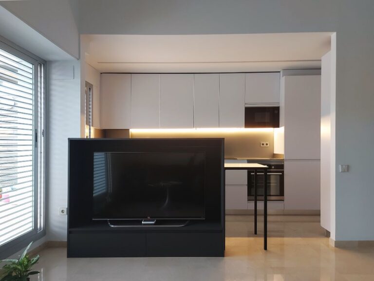 Vista frontal de cocina blanca pequeña abierta al salón con mueble negro para TV que separa la cocina del salón con una mesa detrás