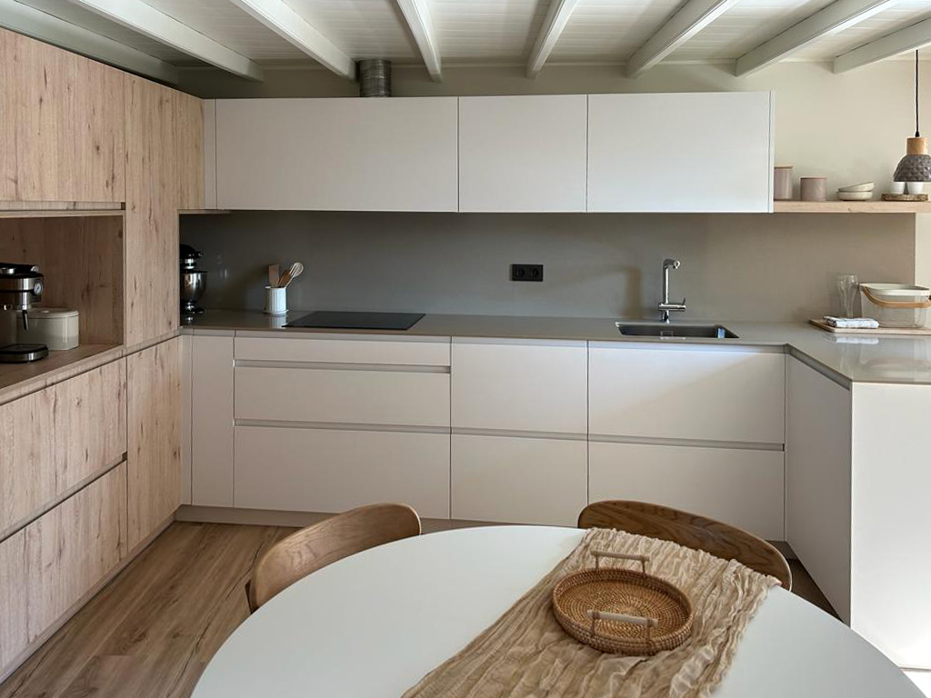 Vista frontal de cocina en ele con muebles en color blanco y en madera, con encimera gris equipada con vitrocerámica y fregadero