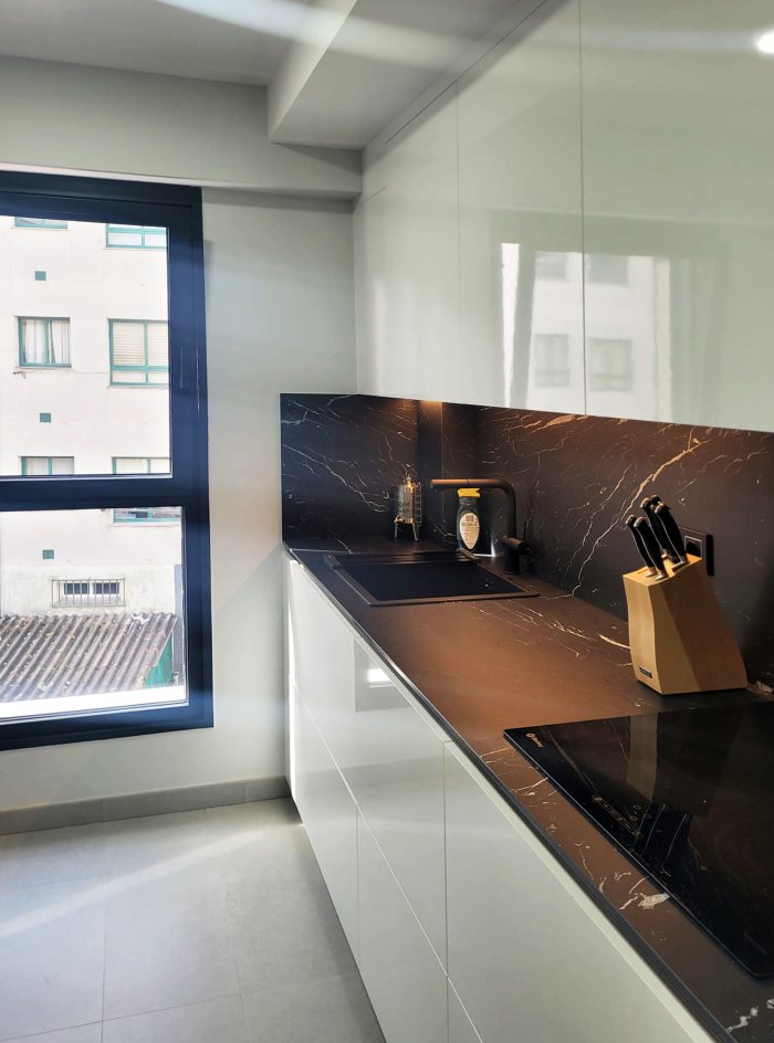 Vista lateral de cocina con muebles en acabado blanco y encimera negra con fregadero negro y vitrocerámica.
