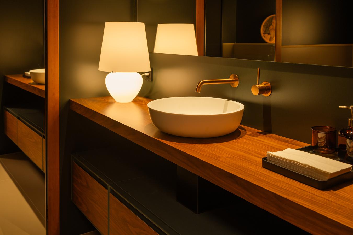 Baño de diseño en la exposición de Kitchen in Santiago, con un elegante mueble de madera, un lavabo redondo blanco y grifería en acabado dorado empotrada en la pared.