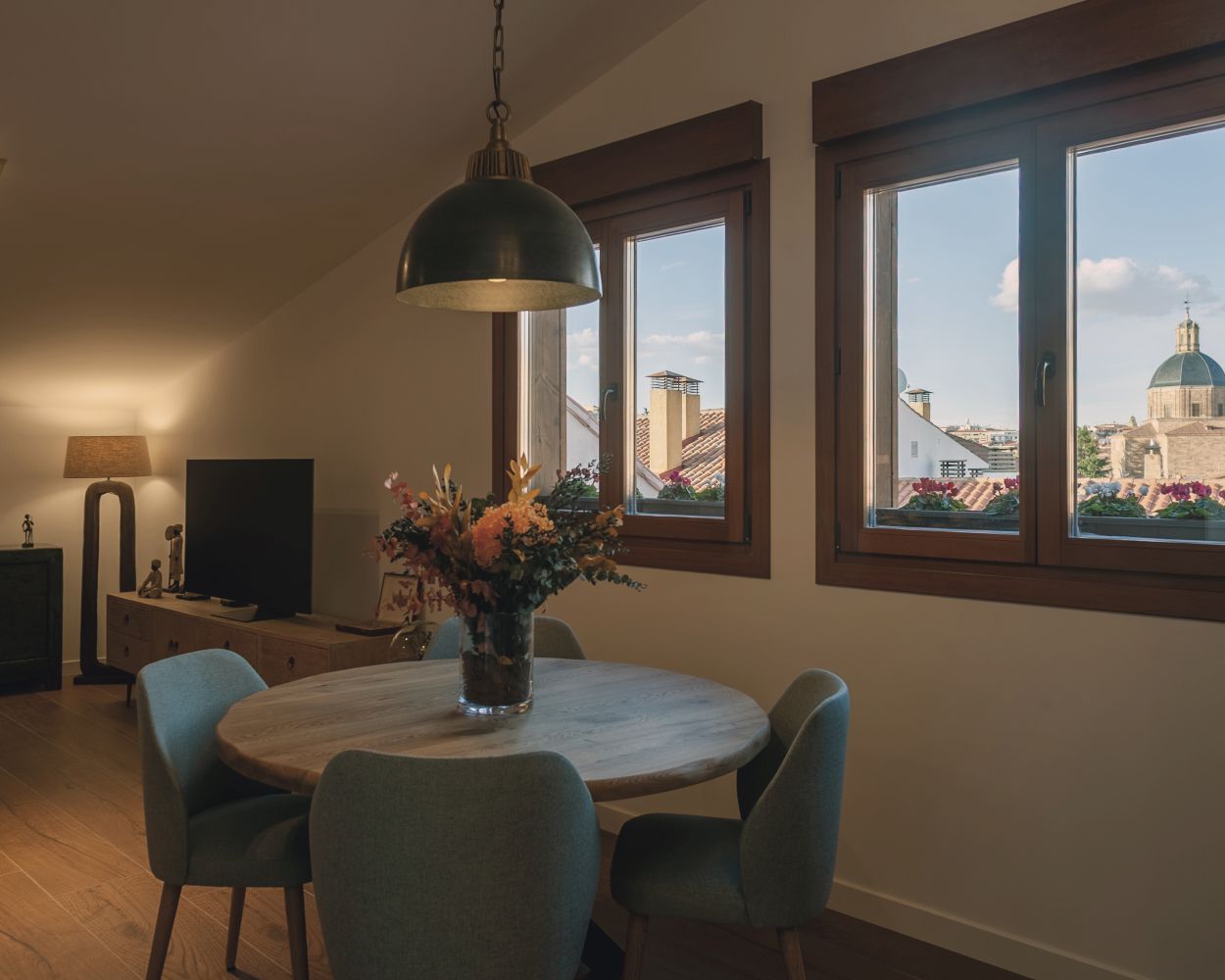 Comedor integrado junto a la cocina con mesa redonda de madera, iluminación colgante y grandes ventanas con vistas a los tejados de la ciudad.