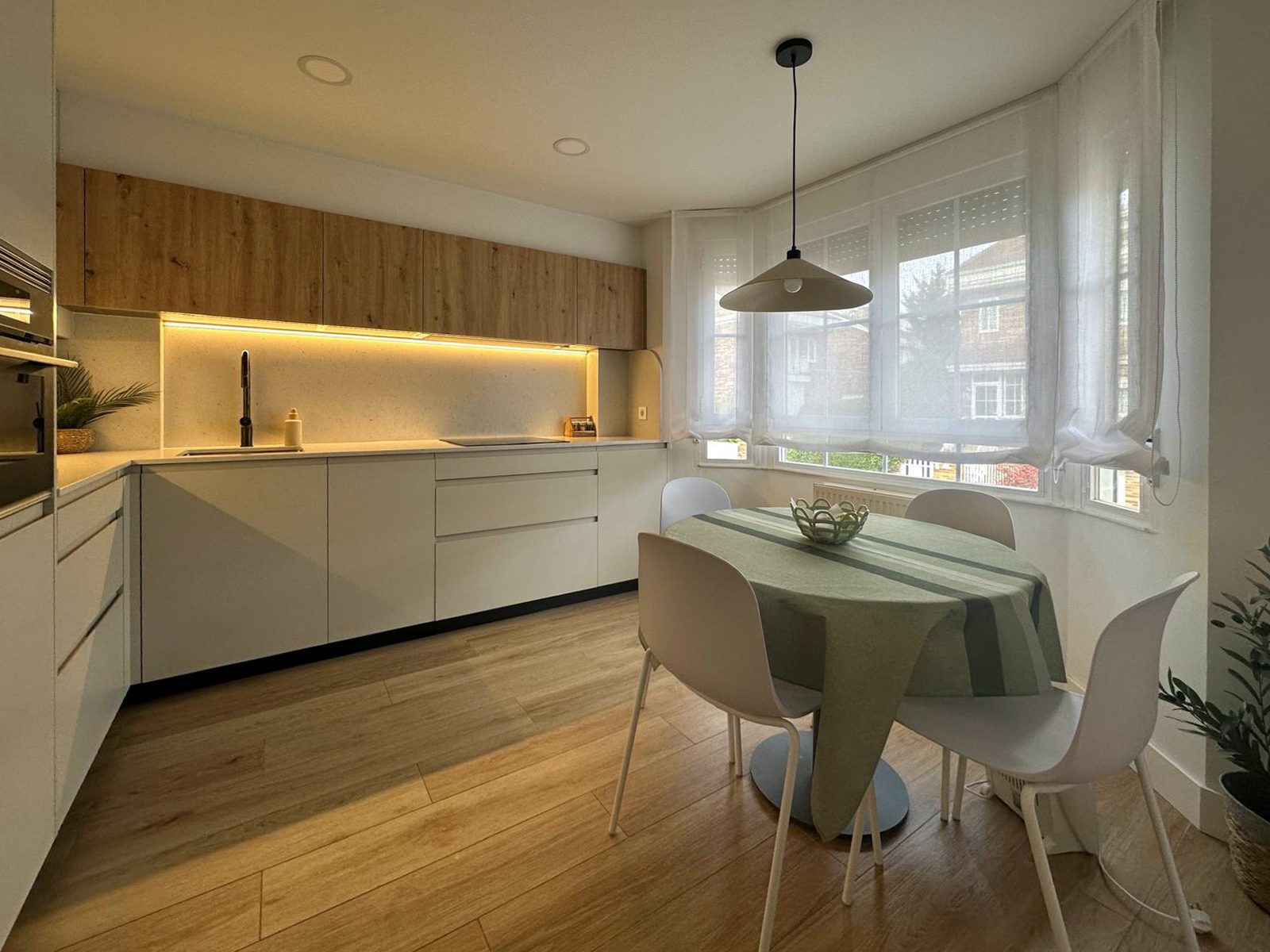 Cocina con iluminación indirecta bajo los muebles altos de Roble Dorado y mesa redonda junto a la ventana