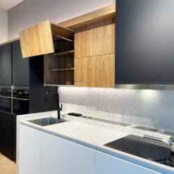 Detalle de muebles en negro, blanco y acabado madera en cocina moderna en Kitchen in Salamanca Vista detallada de la cocina en Kitchen in Salamanca, con mueble alto abierto en madera, muebles bajos en blanco y columnas grises antracita junto a la placa y el fregadero.