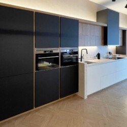 Cocina negra equipada con vitrina y electrodomésticos en Kitchen in Salamanca Vista de cocina en Kitchen in Salamanca, con columnas grises antracita que incorporan electrodomésticos y frigorífico encastrado, muebles bajos en blanco mate con placa y fregadero, altos en madera con iluminación, y vitrina negra mate con cristal al fondo.