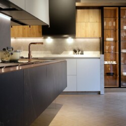 Isla de cocina moderna en cerámica con cocina en un frente en blanco, madera y cristal al fondo Cocina moderna con distribución en isla. Al fondo se ve una gran cocina distribuida en un frente con mobiliario combinado en blanco y madera y muebles columna vitrina