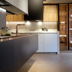 Isla de cocina moderna en cerámica con cocina en un frente en blanco, madera y cristal al fondo Cocina moderna con distribución en isla. Al fondo se ve una gran cocina distribuida en un frente con mobiliario combinado en blanco y madera y muebles columna vitrina