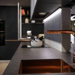 Detalle de grifo y fregadero en acabado cobre dorado Detalle de grifo y fregadero en acabado cobre dorado de cocina en isla negra con composición de mobiliario columna al fondo