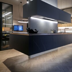 Cocina en isla de diseño moderno en Kitchen in Salamanca Cocina de diseño en isla con muebles en acabados cerámica y gris, con cocina blanca y madera al fondo a la derecha, y armario vestidor con puertas de cristal a la izquierda