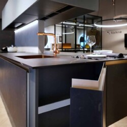 Mueble de apertura lateral modelo FUSSION de Senssia en acabado cerámica KELYA Detalle de mueble de apertura lateral abierto en isla de cocina negra con grifo dorado