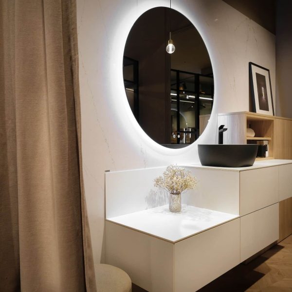 Composición moderna de baño con muebles bajos en color blanco y columna en color abeto natural. Dispone de un gran espejo circular y el lavabo es sobreencimera
