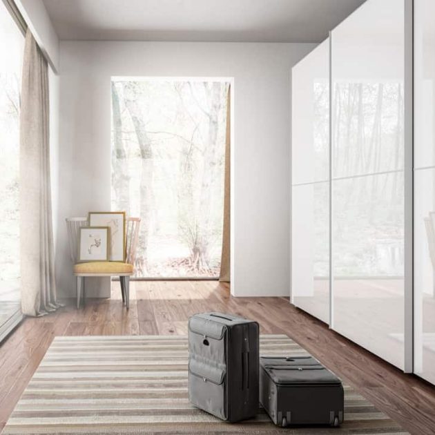 Vista lateral de armario amplio en color blanco de aluminio y cristal en habitación luminosa con alfombra, maletas y silla con cuadros