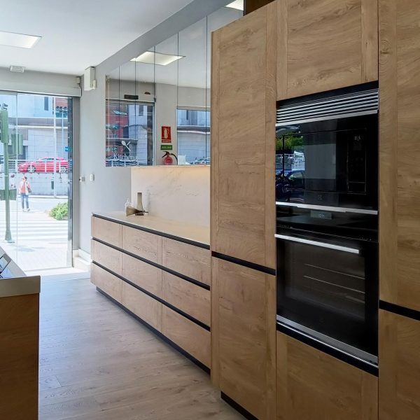 Vista lateral de exposición en tienda con cocina moderna de un frente en acabado madera con horno y microondas integrados. Al fondo, está la puerta de entrada a la tienda