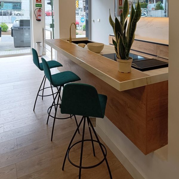 Detalle de isla de cocina apoyada en bancada de obra con espacio para 3 comensales y taburetes en tono verde. Incluye fregadero y placa de cocina.