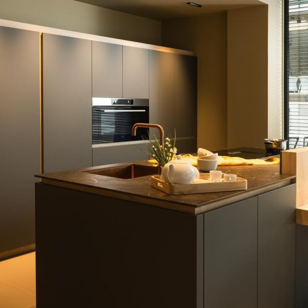 Cocina moderna con muebles en Verde Comodoro, isla central con encimera oscura, grifo dorado, y tabla para comidas en exposición de tienda Kitchen in Santiago.