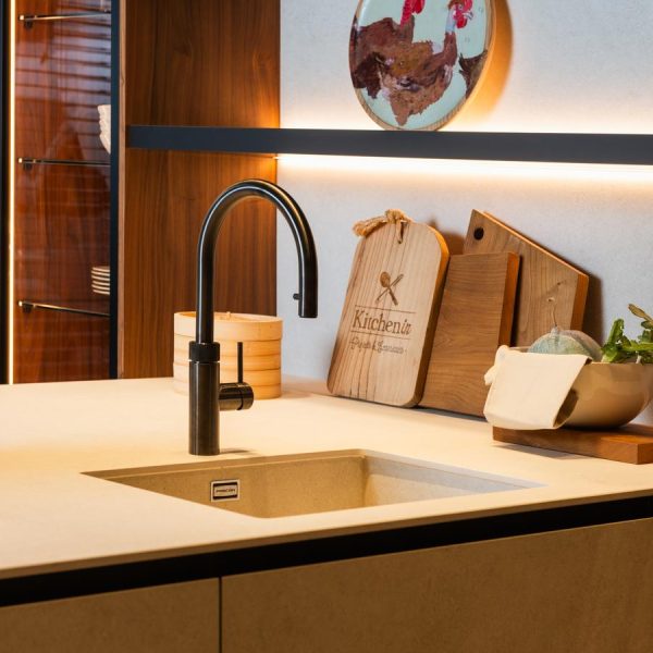 Detalle de península de una cocina moderna en exposición de Kitchen in Santiago, con muebles de madera natural y acabados en negro mate y tres columnas vitrina iluminadas
