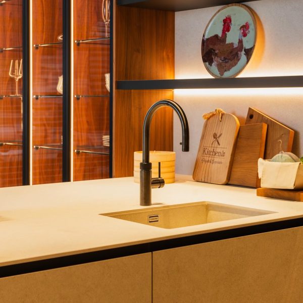 Detalle de península de una cocina moderna en exposición de Kitchen in Santiago, con muebles de madera natural y acabados en negro mate y tres columnas vitrina iluminadas