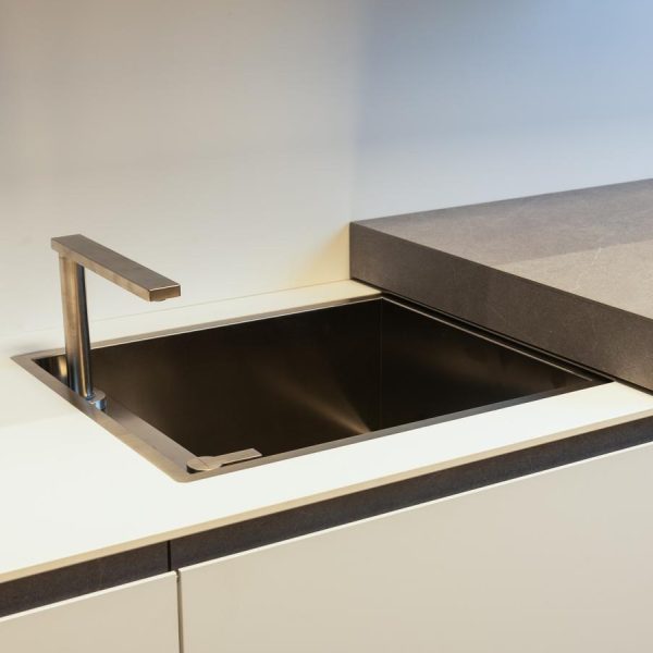 Fregadero integrado con grifo escamoteable, oculto bajo una encimera deslizante de compacto en Blanco Fénix en exposición de la tienda Kitchen in Santiago.