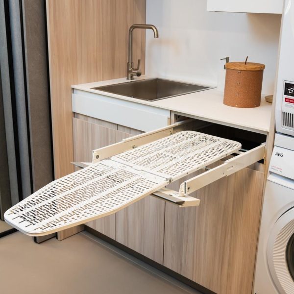 Lavandería moderna en la exposición de la tienda Kitchen in Santiago con bajo fregadero en acabado Maple Gentle y una mesa de planchar extraíble integrada.