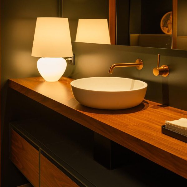 Baño de diseño en la exposición de Kitchen in Santiago, con un elegante mueble de madera, un lavabo redondo blanco y grifería en acabado dorado empotrada en la pared.