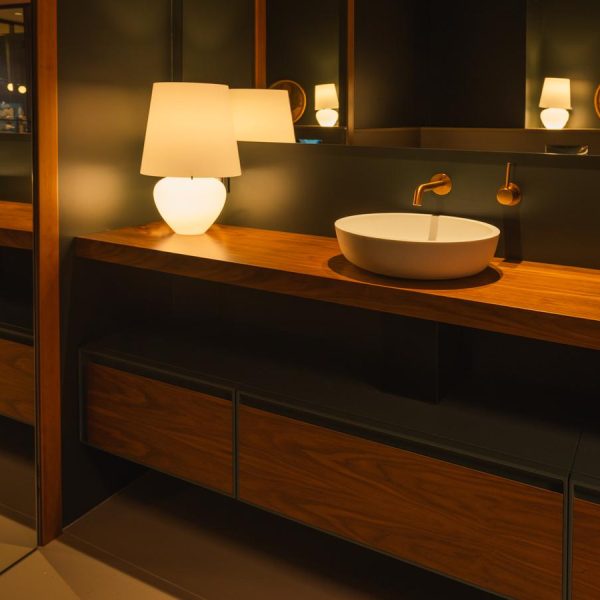 Baño de diseño en la exposición de la tienda Kitchen in Santiago, con una encimera de madera natural, un lavabo redondo blanco, grifería dorada empotrada y armario de la marca EMEDE