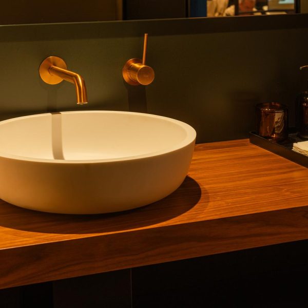 Lavabo de cerámica blanca sobre una encimera de madera natural, acompañado de una grifería empotrada en acabado dorado y una bandeja con accesorios de baño