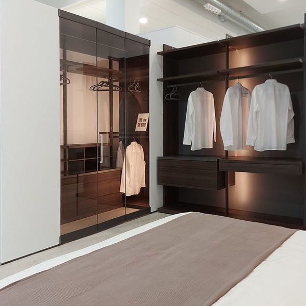 Vista desde la cama de dos armarios vestidores en forma de ele en dormitorio moderno en acabado madera y uno con puertas de cristal