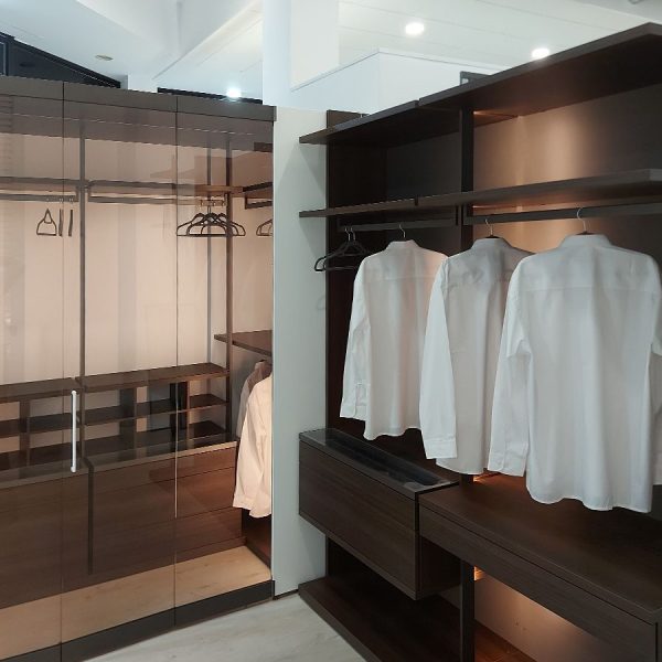 Vista lateral de armario de cristal con puertas en fuelle y vestidor abierto en madera oscura con camisas colgadas