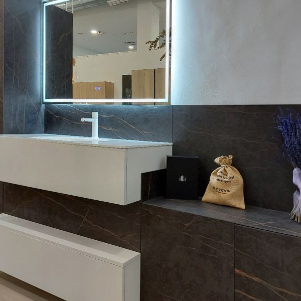 Vista lateral de composición moderna para baño en exposición con lavabo en blanco, muebles en acabado cerámica oscura y espejo rectangular con leds