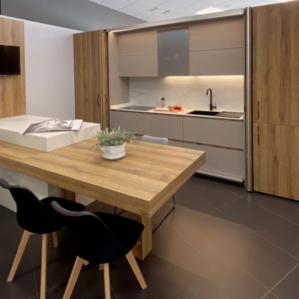 Vista lateral de cocina en blanco y madera con península con mesa integrada para comidas y TV sobre pared