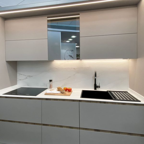 Vista frontal de cocina blanca pequeña con vitrocerámica y fregadero negro integrados, espejo entre los muebles altos y luz led bajo estos